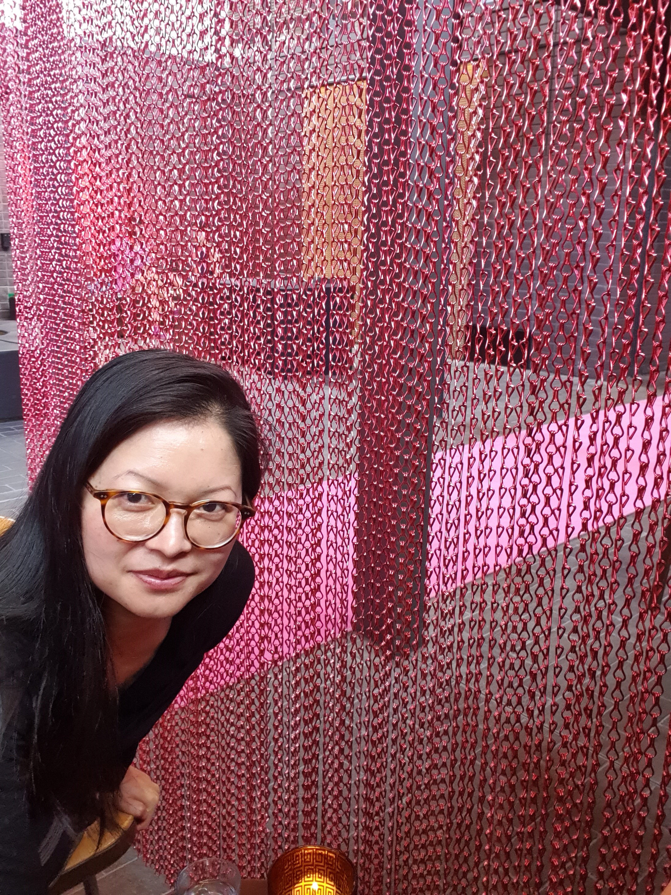 Photograph of a person with long black hair and glasses leaning into the picture in front of a pink art installation